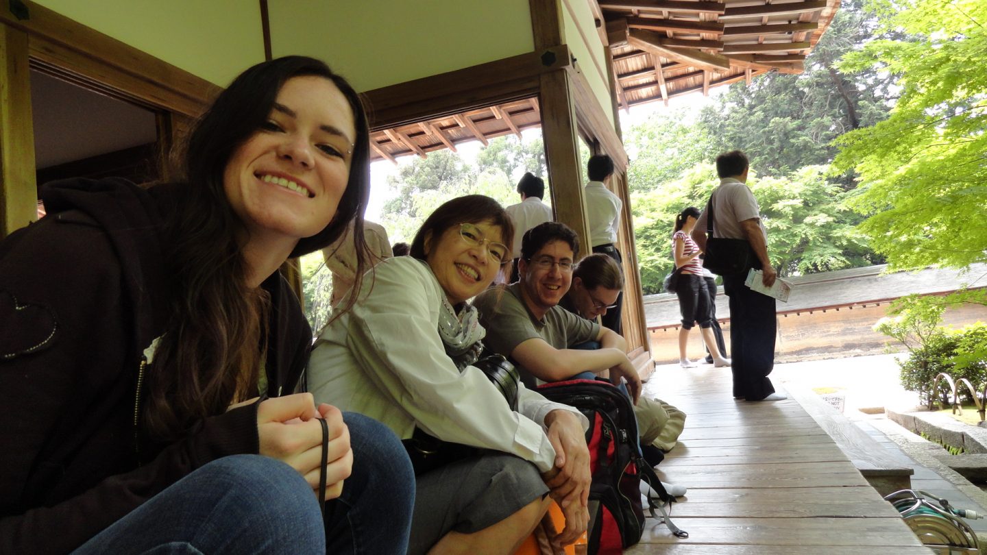 Four people sit in a row on the outdoor porch of a wooden house. Three of them are looking at the camera and smiling, the last person in the row is looking down. There are several other people in the background of the photo, facing away from the camera