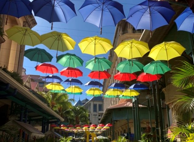 Shot of a street with dozens of brightly colored umbrellas hanging above. The Umbrellas are yellow, blue, green and red. The sky is sunny and bright blue and there are palm trees in the background