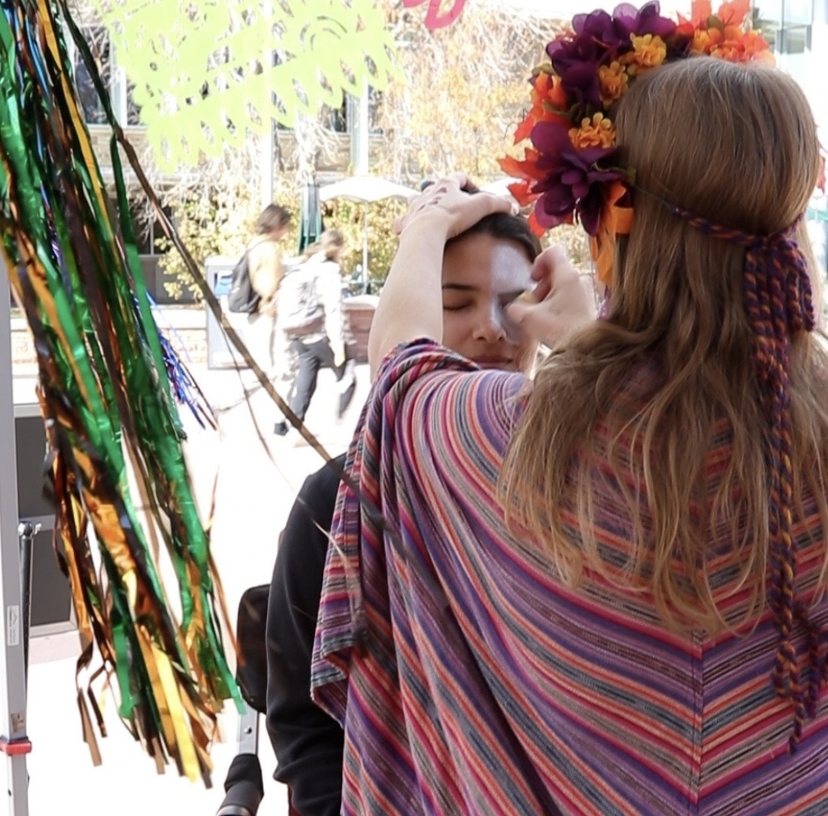 Person with long hair with flowers is applying white makeup to another person