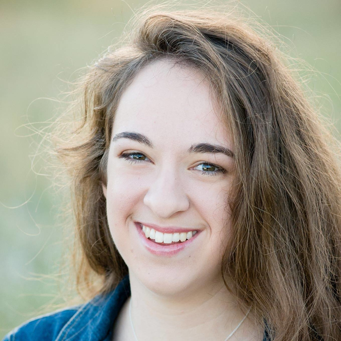 A close-up portrait of a person with shoulder-length brown hair smiling warmly at the camera. They are wearing a blue top and photographed against a soft, blurred green background.